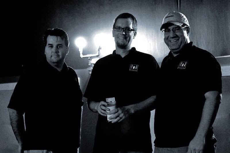 Three men in matching black shirts with logos, standing together in a dimly lit room.