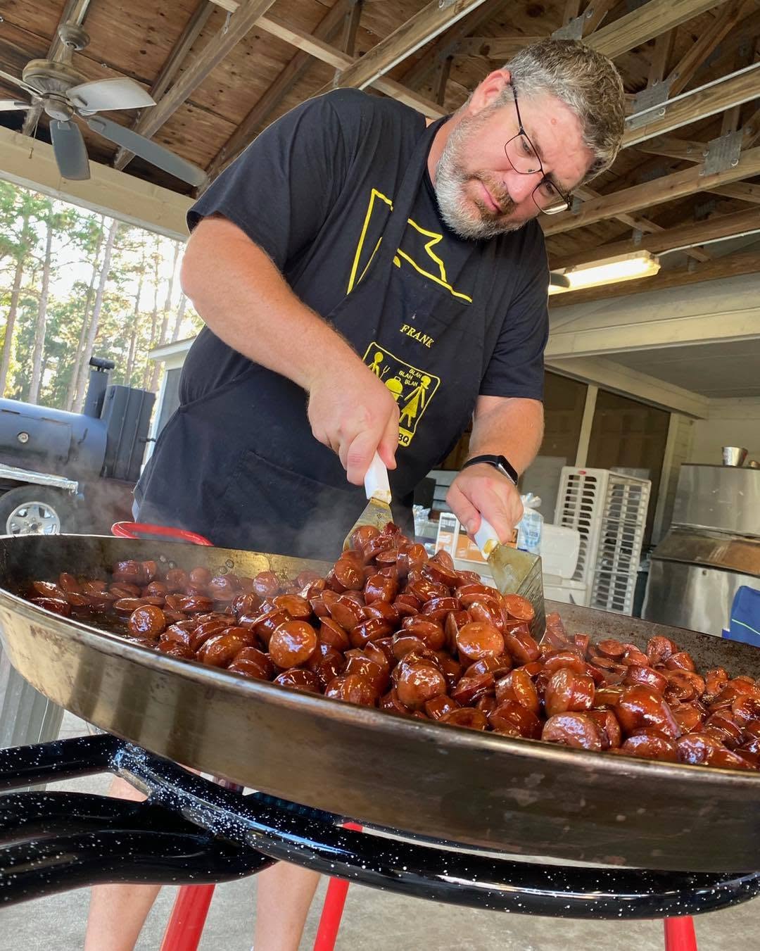 A man wearing a black graphic t-shirt and glasses is cooking a large batch of glazed sausage in a massive stainless steel pan on a barbecue grill stand inside a covered outdoor kitchen area.