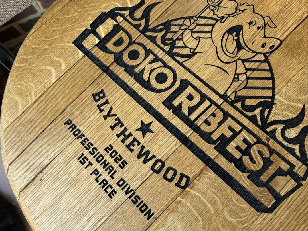 Close-up of a round wooden surface engraved with a black logo reading 'Doko Ribfest' and 'Blythewood 2025 Professional Division 1st Place,' plus a pig illustration and flames.